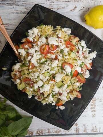 feta and orzo pasta salad in a black bowl with some lemons on the side