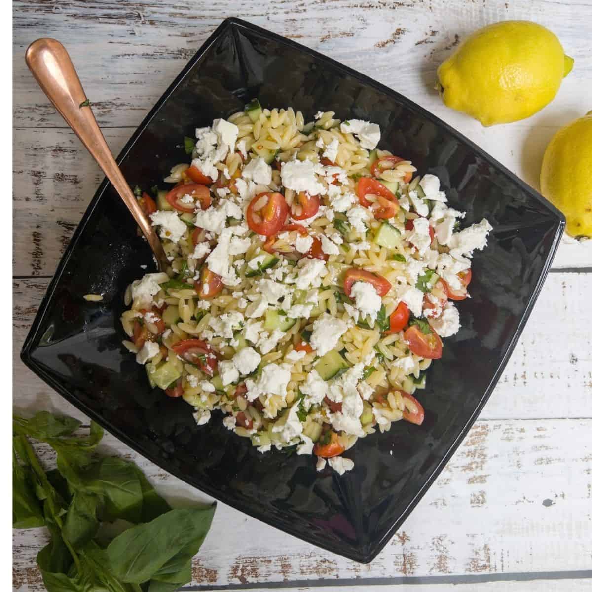 feta and orzo pasta salad in a black bowl with some lemons on the side