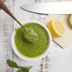 green basil pesto in a white bowl with a spoon in it and lemon on a chopping board.