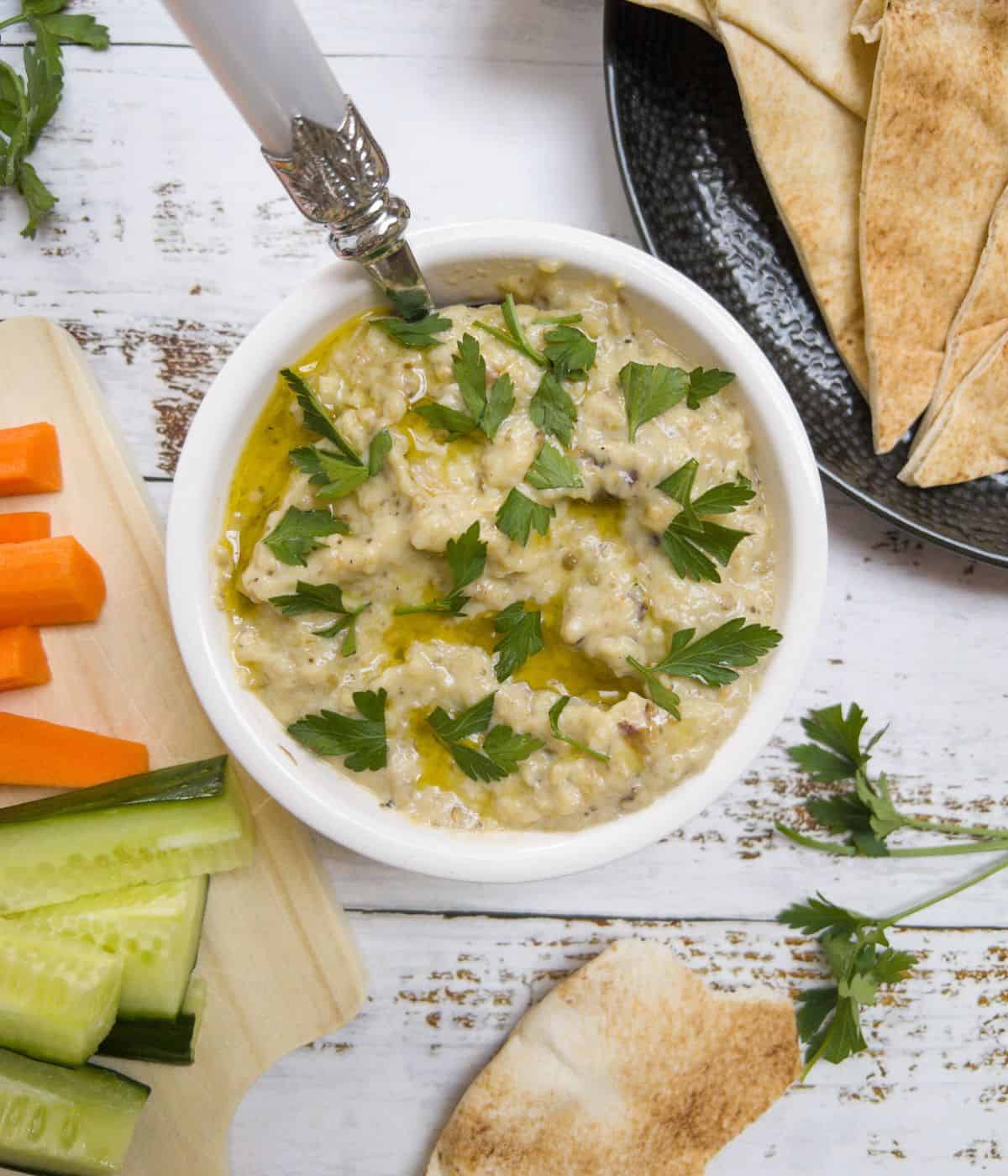 a small white bowl filled with baba ganoush with some flatbread and crudities on the side.