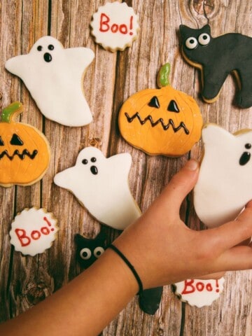 Selection of halloween cookies including ghosts, pumpkins and black cats, with a hand picking up the ghost cookie.