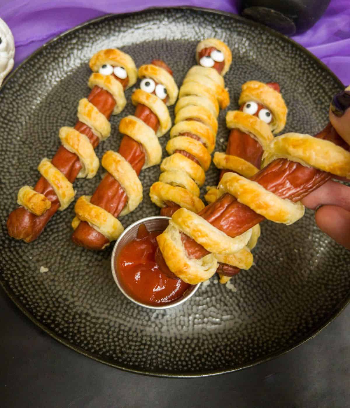 sausage mummies (frankfurters wrapped in puff pastry strips) being dipped in a pot of ketchup