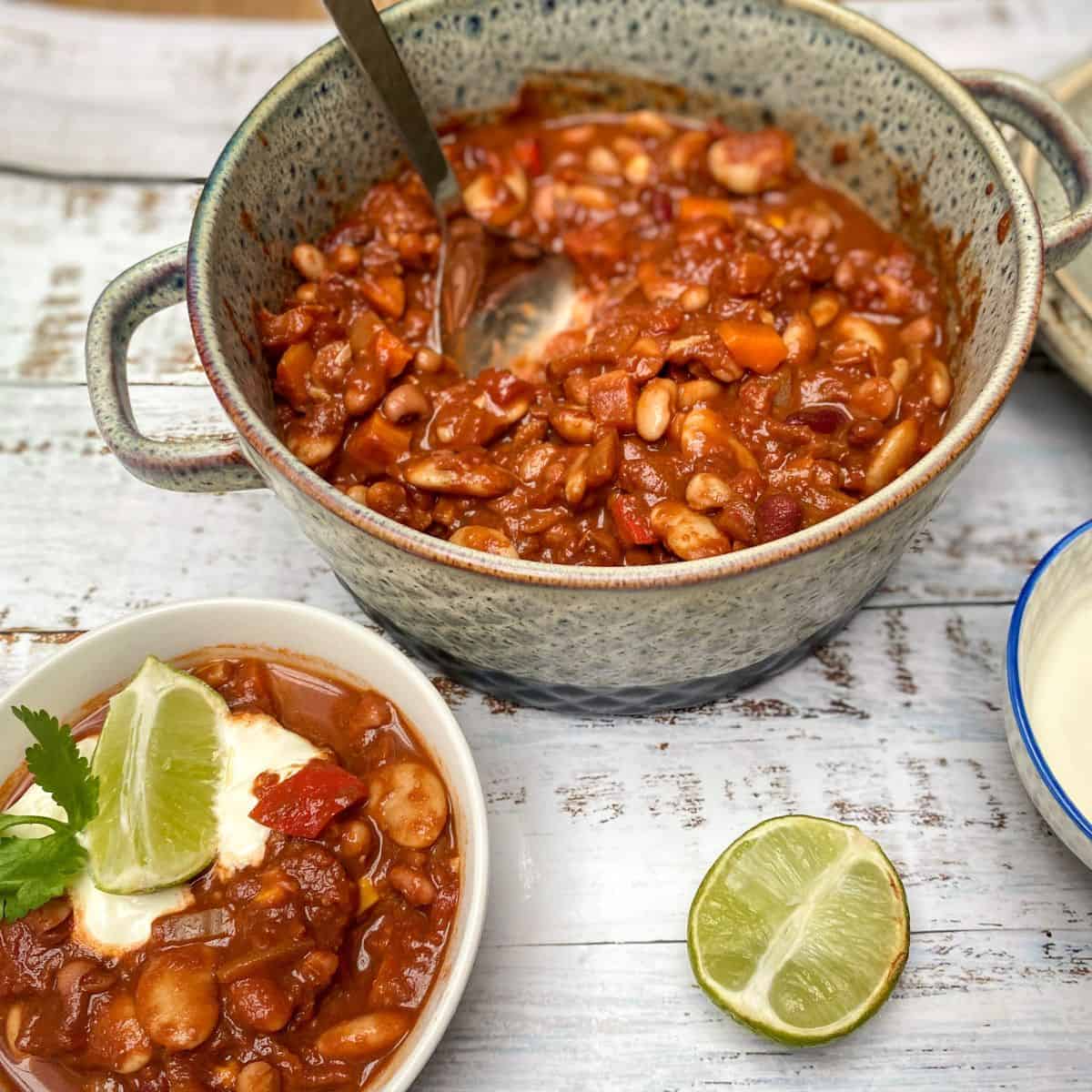 mixed bean chilli in a casserole dish with a bowl of chilli next to it garnished with a lime and sour cream.