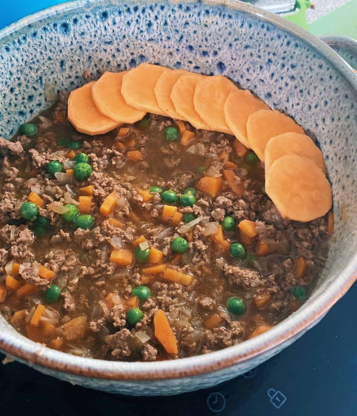 cooked lamb mince carrot and pea mixture being topped with sliced sweet potato