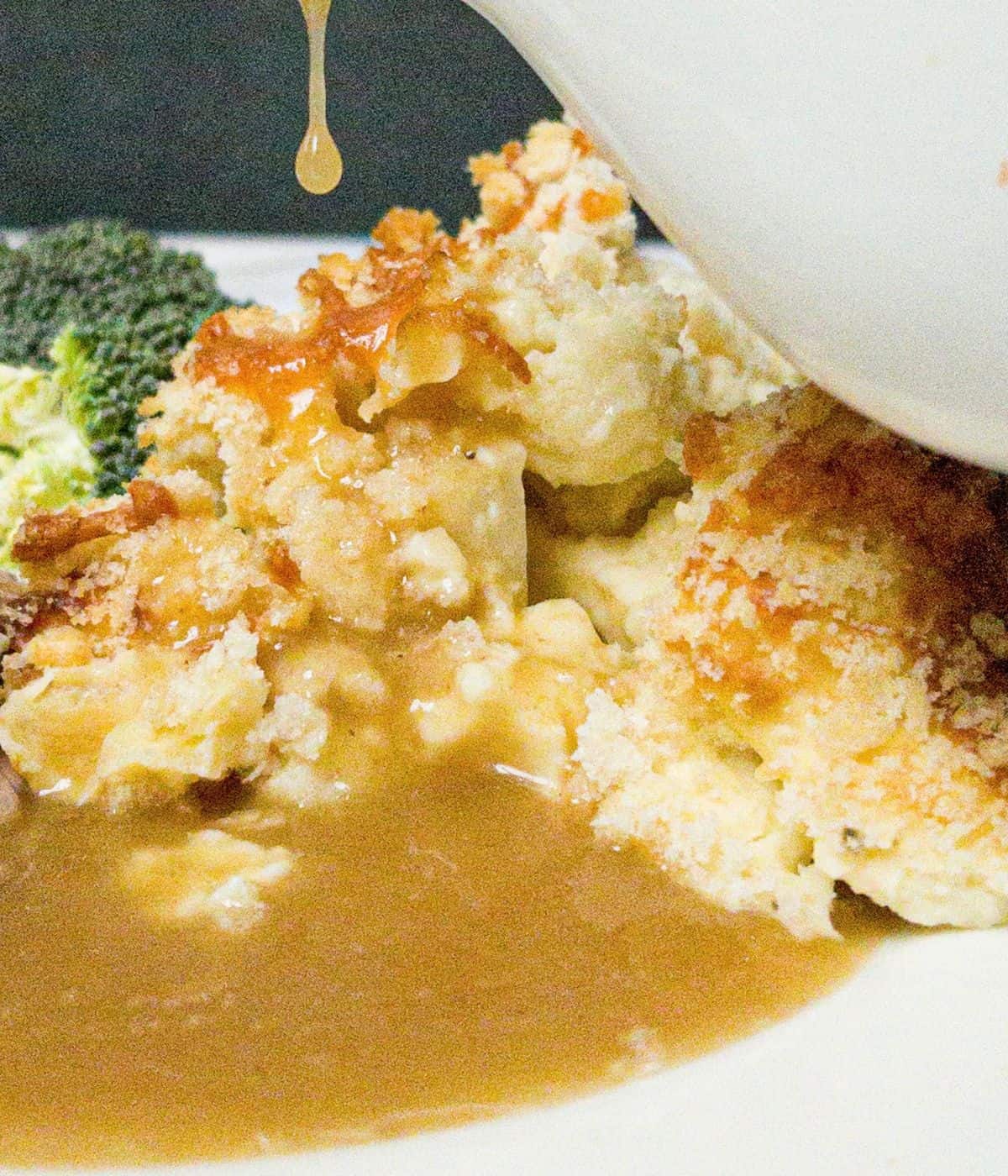 A white plate with cauliflower cheese and broccoli on it. A gravy dish is pouring gravy onto the plate.
