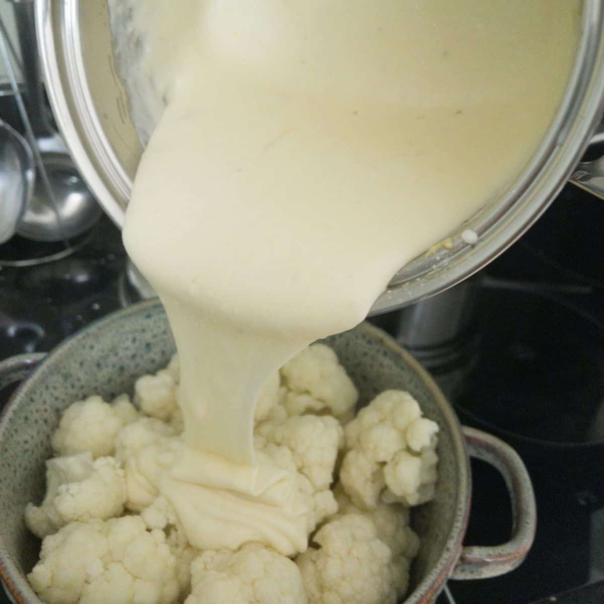 Cheese sauce being poured from a saucepan into a casserole dish full of cauliflower.