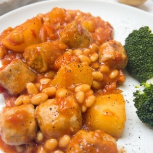 close up photo of sausage and baked bean casserole on a white plate with some broccoli.
