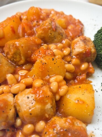 close up photo of sausage and baked bean casserole on a white plate with some broccoli.