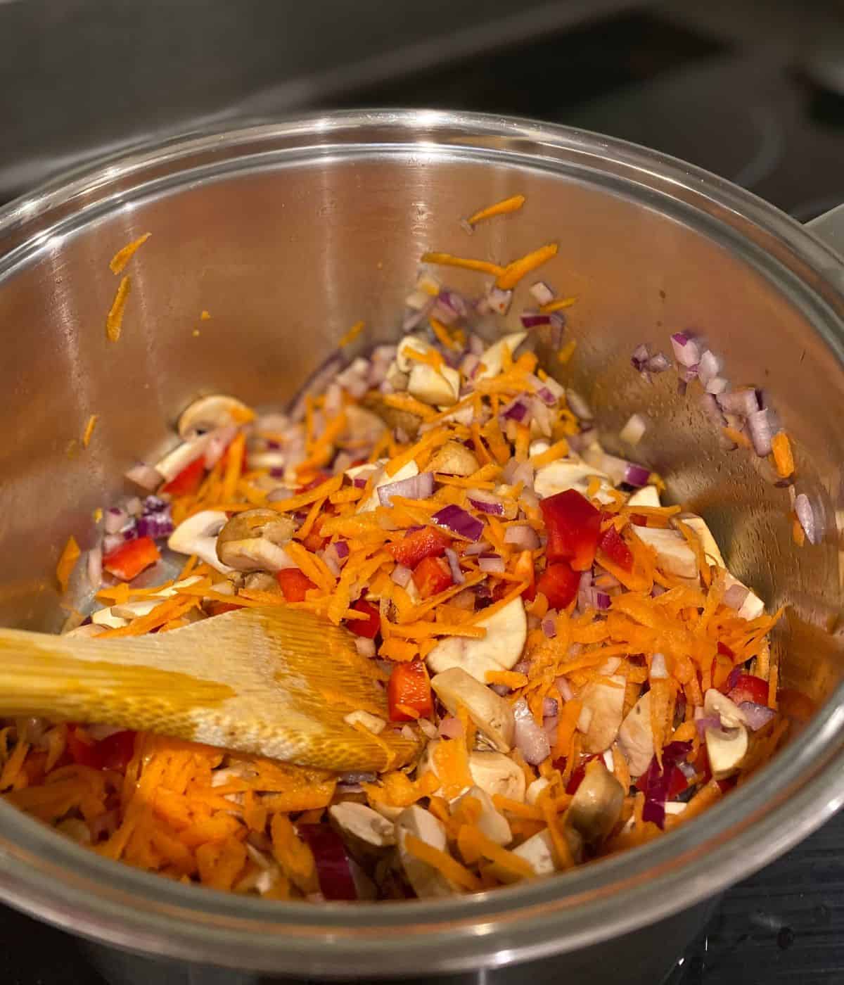 carrot, mushroom, onion and peppers sweating in a saucepan, being stirred with a wooden spoon.