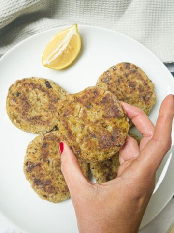A hand picking up a bubble and squeak patty from a plate full of them. Plate is garnished with a slice of lemon.