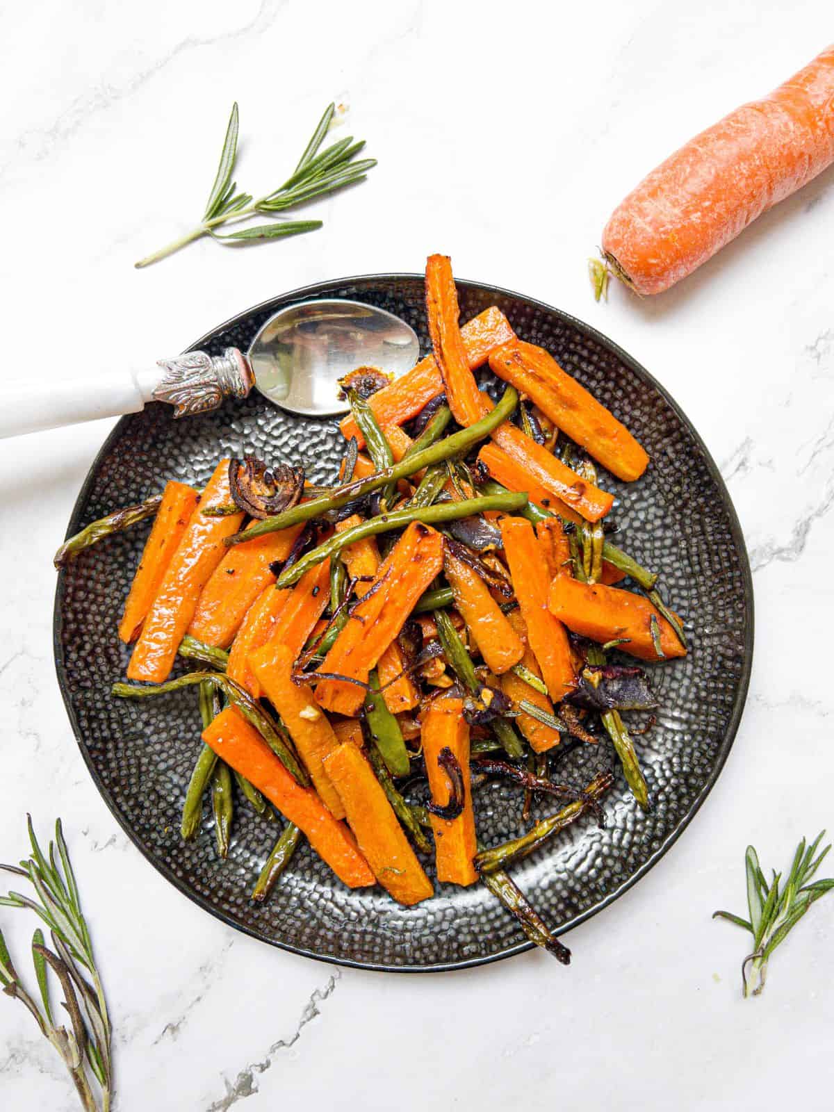 Roasted carrots and green beans on a black plate with a rosemary and carrot garnish.