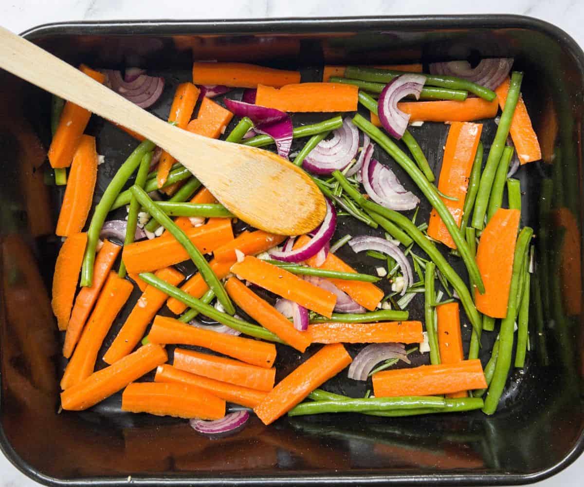 raw carrots, green beans and red onion in a baking tray with a wooden spoon in the tray.