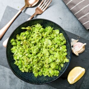 Minted mushy peas in a black bowl on a grey slate with a fork and spoon, garlic and lemon on it .