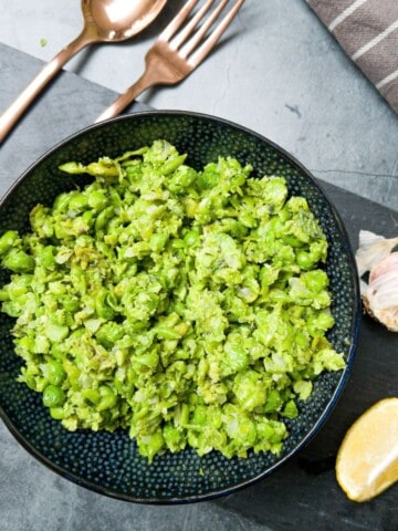 Minted mushy peas in a black bowl on a grey slate with a fork and spoon, garlic and lemon on it .