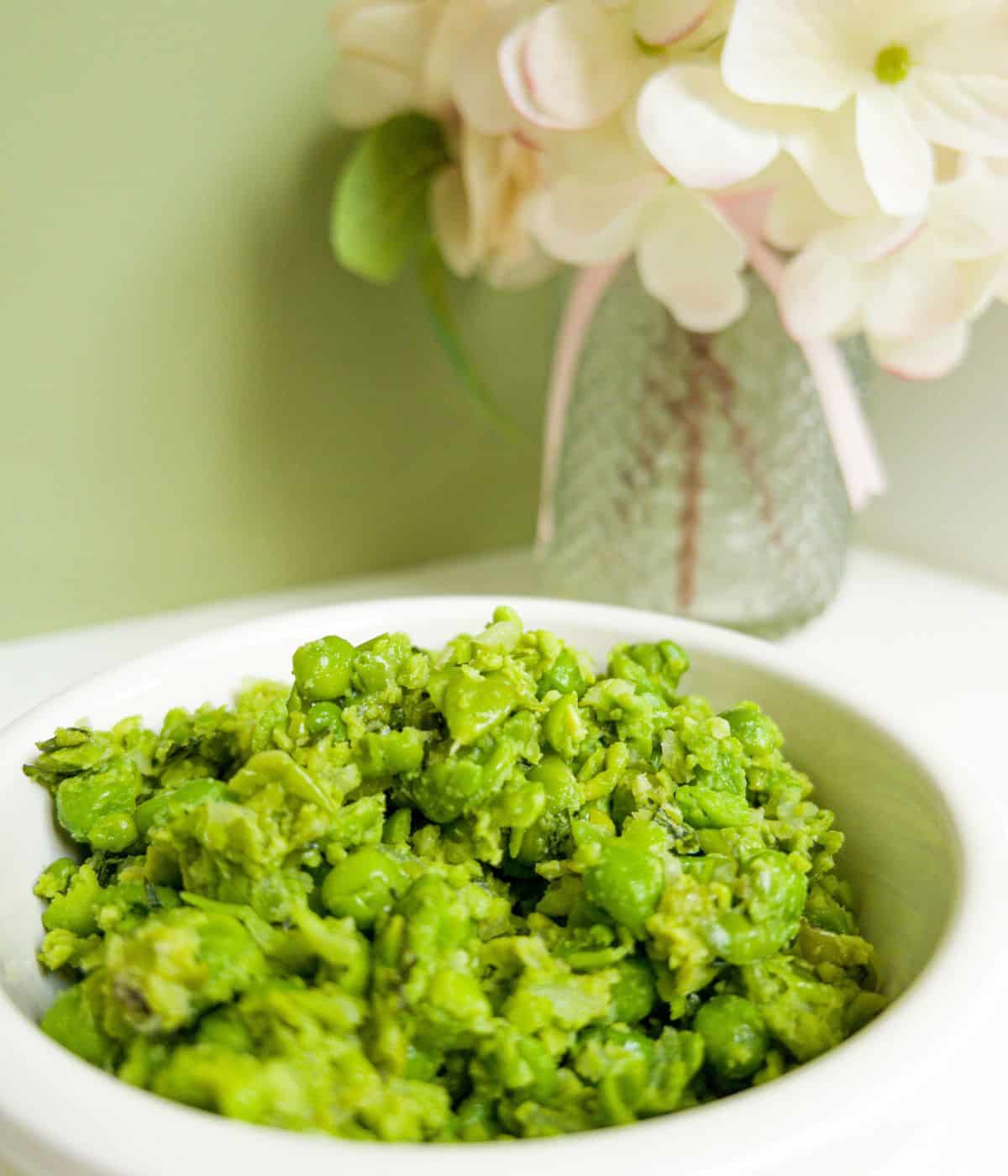 Minted mushy peas in a white bowl with a vase of flowers in the background.