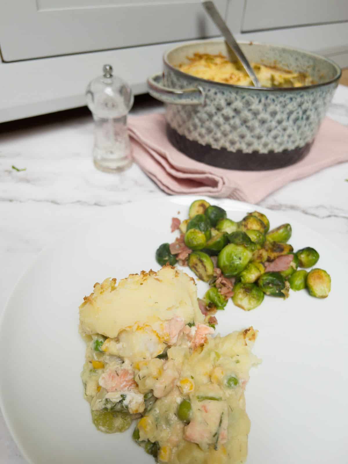 Fish pie and brussel sprouts on a white plate with a salt grinder and pot of fish pie in the background.
