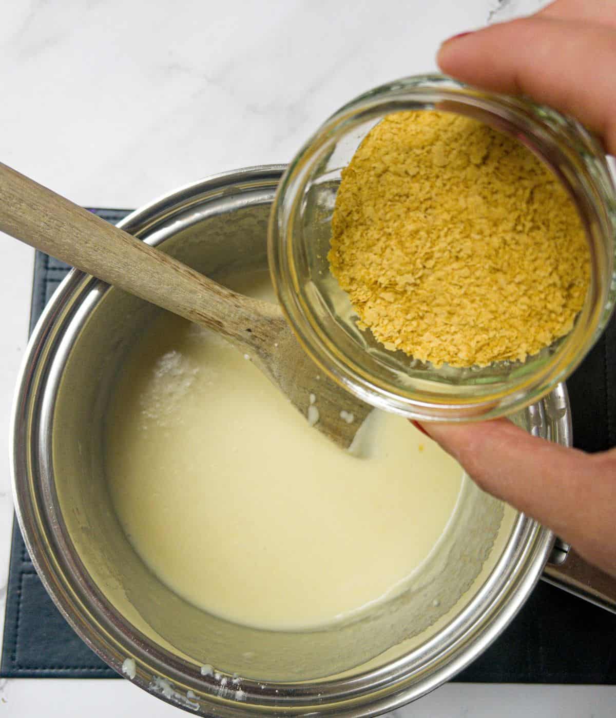 Nutritional yeast being poured into a white sauce in a saucepan.