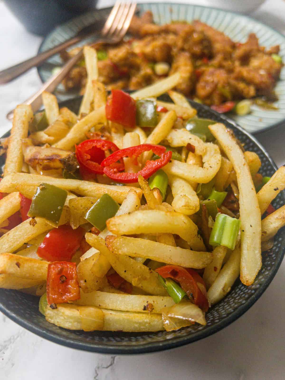 a bowl of fries garnished with chilli, red and green pepper, with a chinese crispy chilli dish in the background.