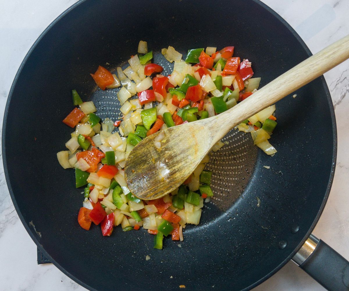 red and green pepper and onion frying in a wok, being mixed by a wooden spoon.