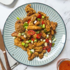 a plate of crispy chicken pieces garnished with spring onion & chilli, with a knife and fork next to the plate and a pot of sweet chilli sauce/