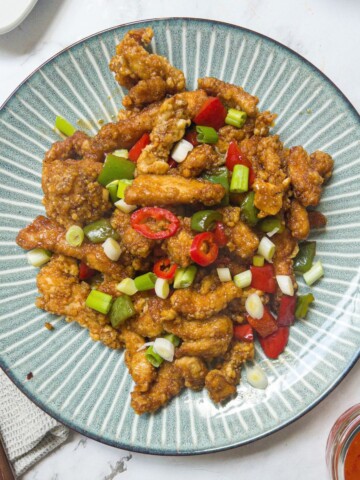 a plate of crispy chicken pieces garnished with spring onion & chilli, with a knife and fork next to the plate and a pot of sweet chilli sauce/
