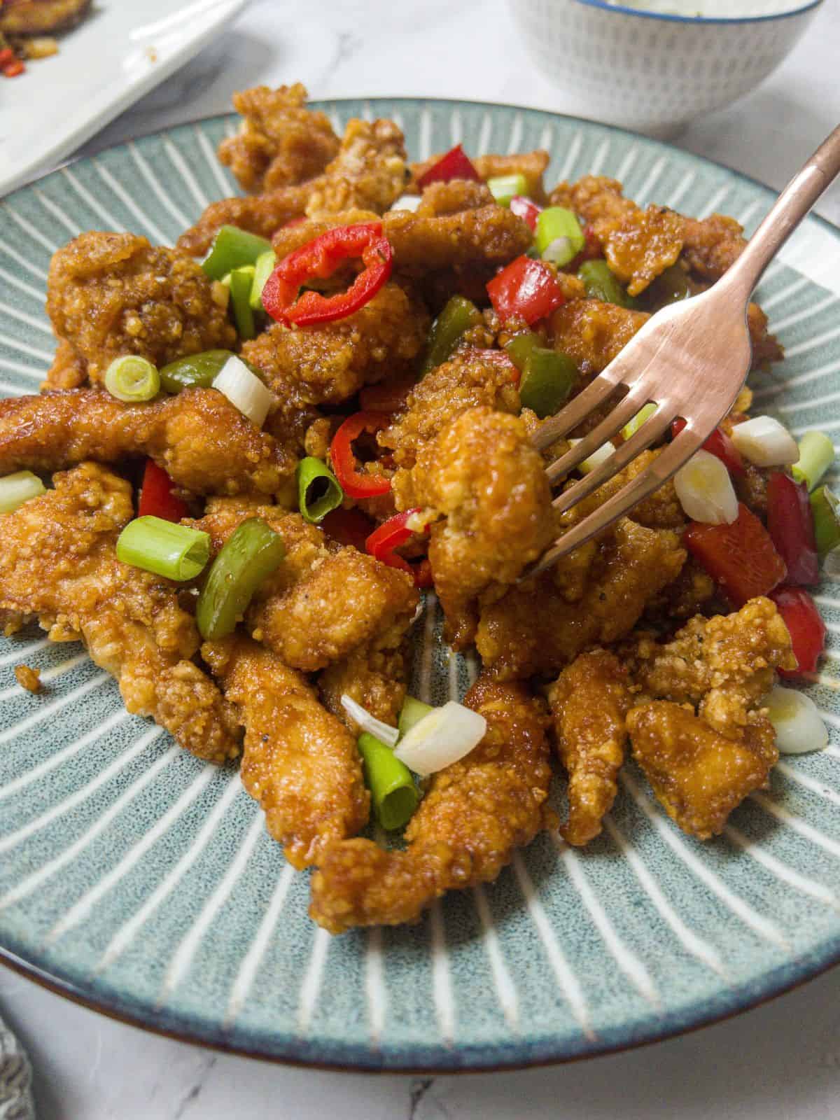 Close up shot of a fork holding a piece of crispy chicken over a plate of crispy chicken, garnished with spring onion and chilli.