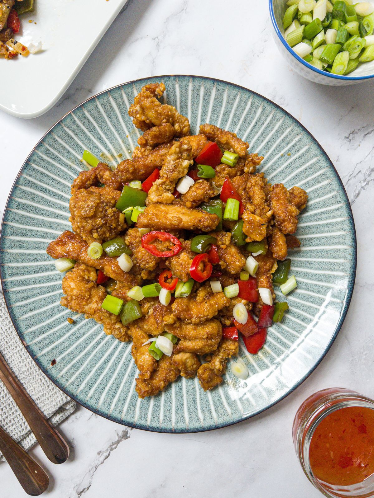 a plate of crispy chicken pieces garnished with spring onion & chilli, with a knife and fork next to the plate and a pot of sweet chilli sauce and spring onions./