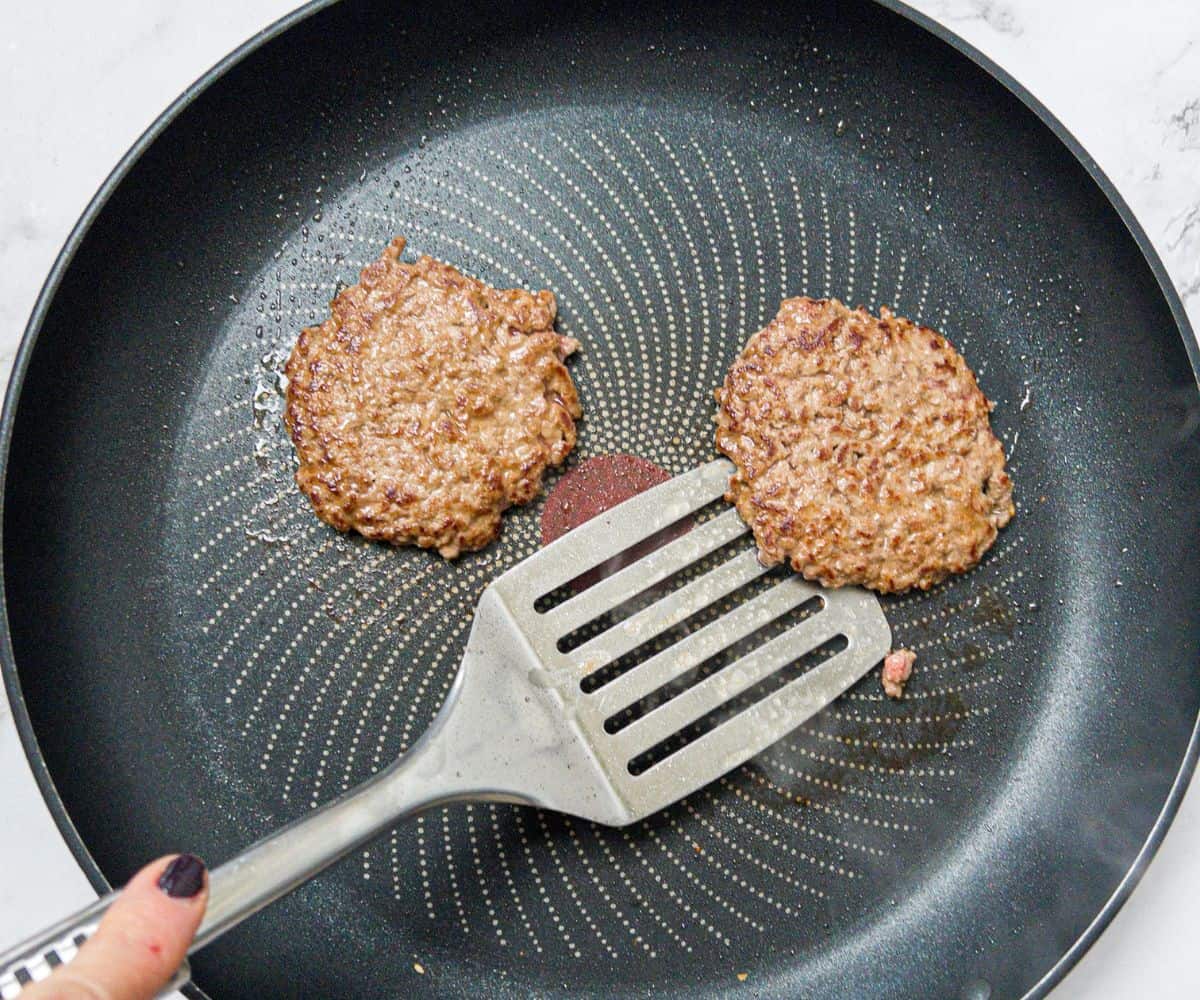 Two burger patties frying in a pan being turned with a spatula.