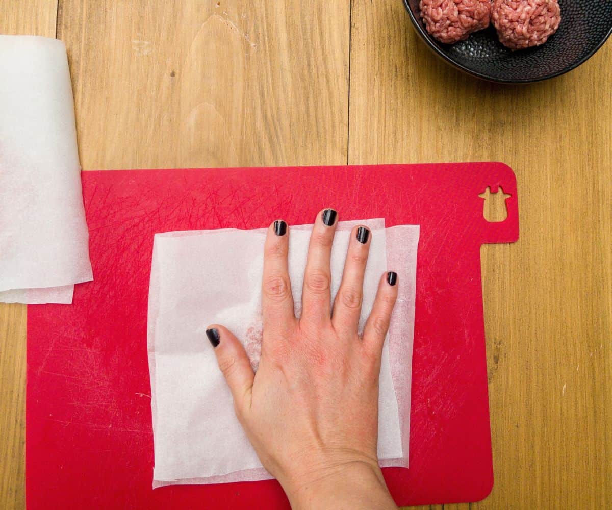 Steak mince being pressed by hand between two sheets of baking paper on a red chopping board.