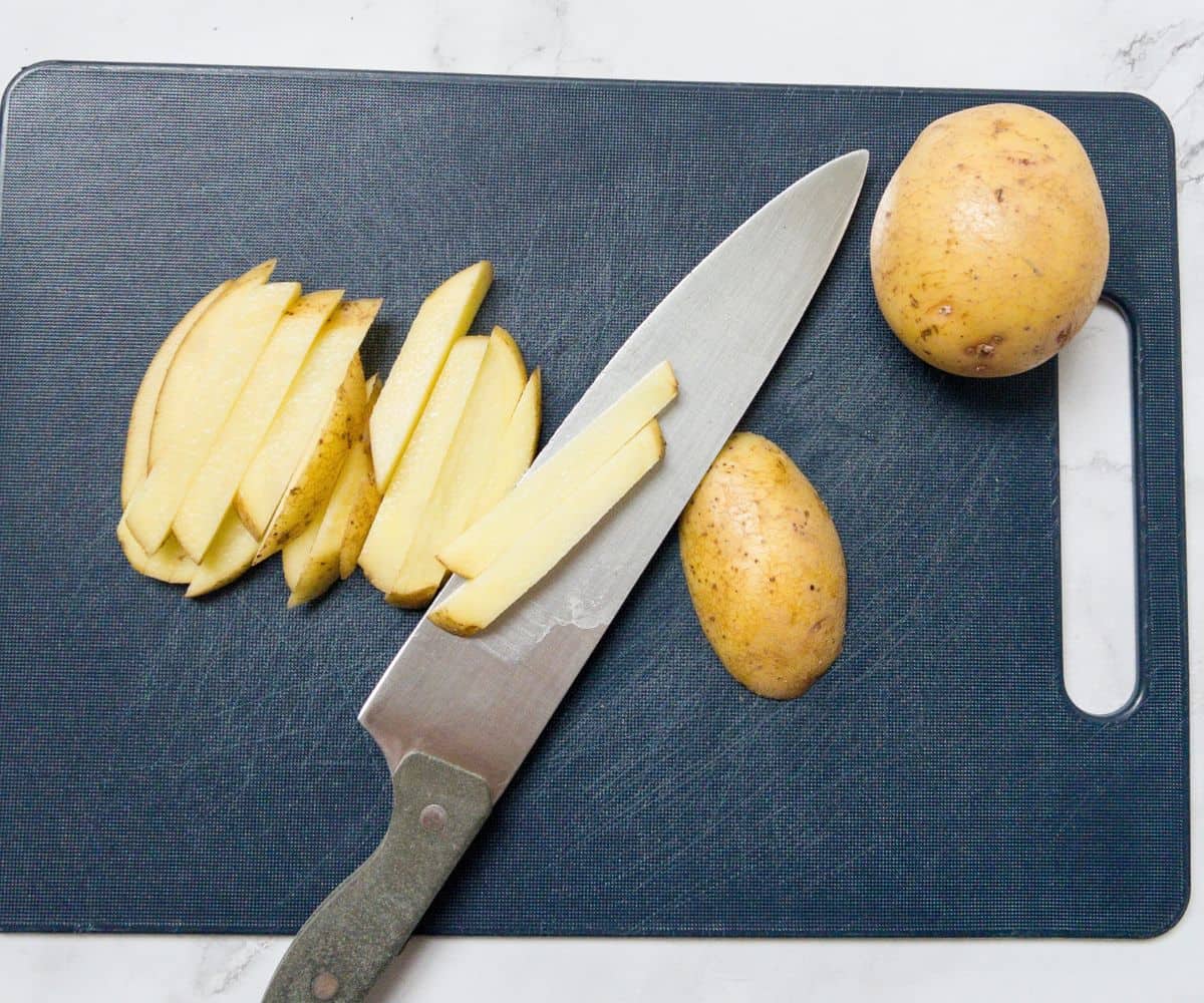 Potatoes being cut into fries on a chopping board with a knife.