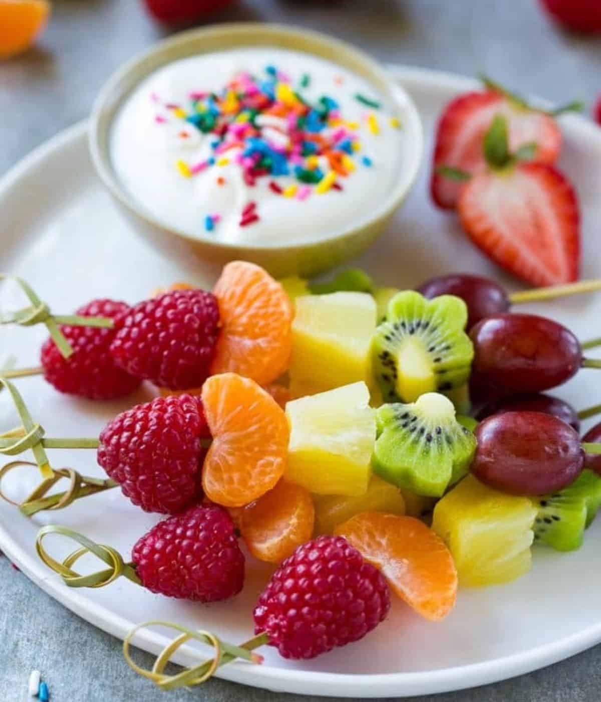Raspberry, satsuma, pineapple, kiwi and grape on a kebab stick, on a plate with a dip with sprinkles and cut strawberry.