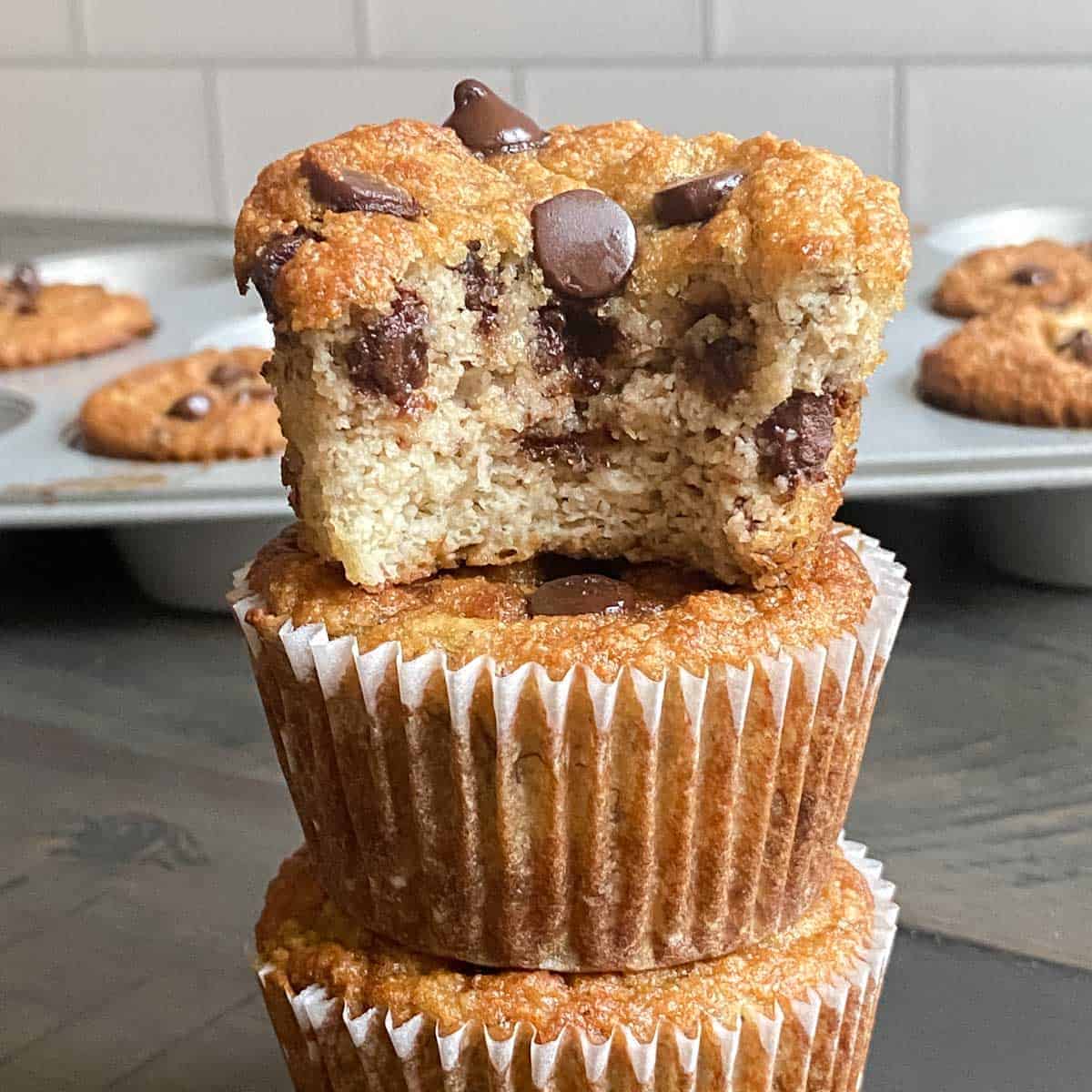 three chocolate chip muffins stacked on top of each other with a bite taken out of the top one. Tray of muffins in the back ground.