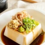 block of tofu topped with spring onion sitting in a white bowl drizzled with soy sauce.