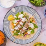 Lamb kofta kebab on a blue plate with a wedge of lemon and toppings in bowls in the background.