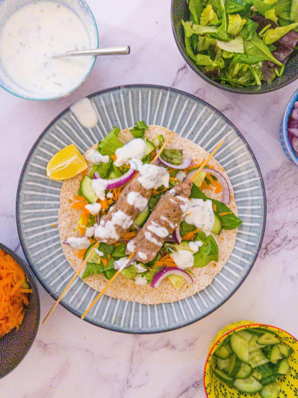 Lamb kofta kebab on a blue plate with a wedge of lemon and toppings in bowls in the background.
