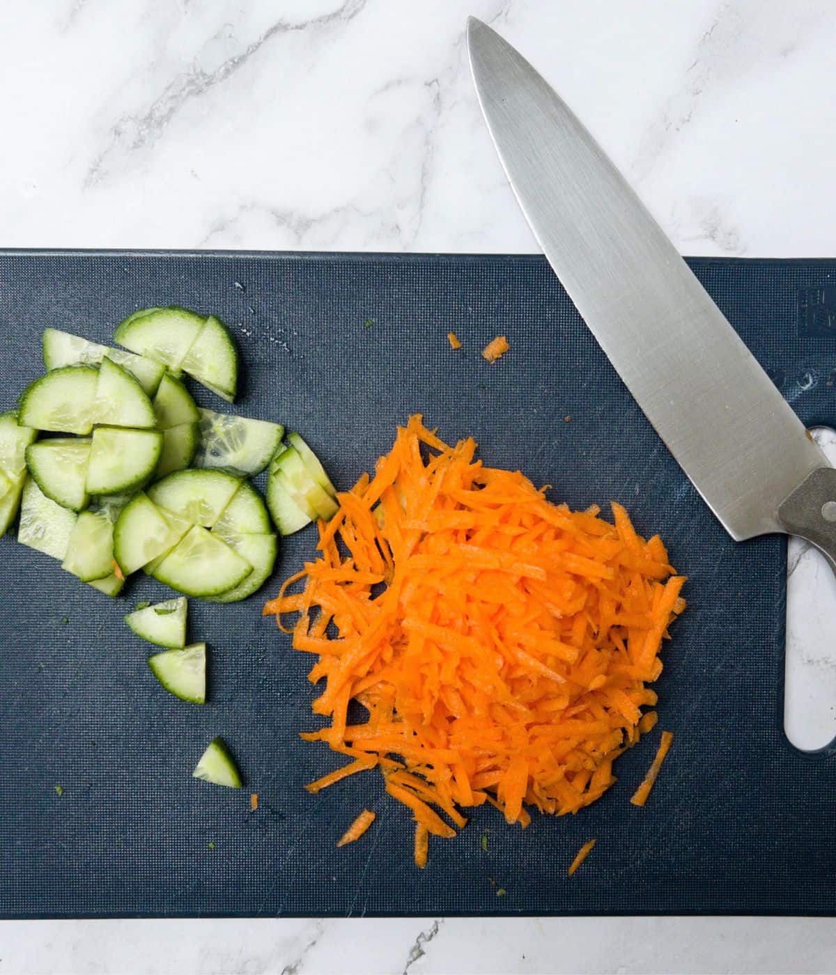 Chopped cucumber and grated carrot on a chopping board with a knife.