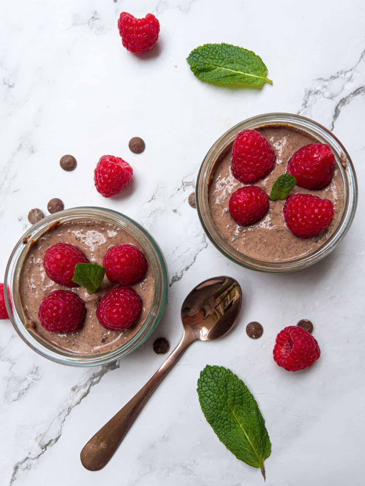 Two pots of chocolate mousse topped with raspberries with mint and raspberries scattered around background.