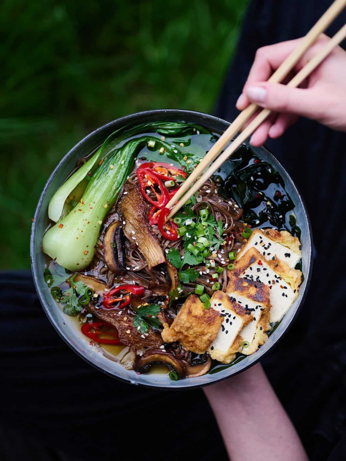 bowl containing tofu, pak choi, chillis in a ramen with a person picking some up with chopsticks.