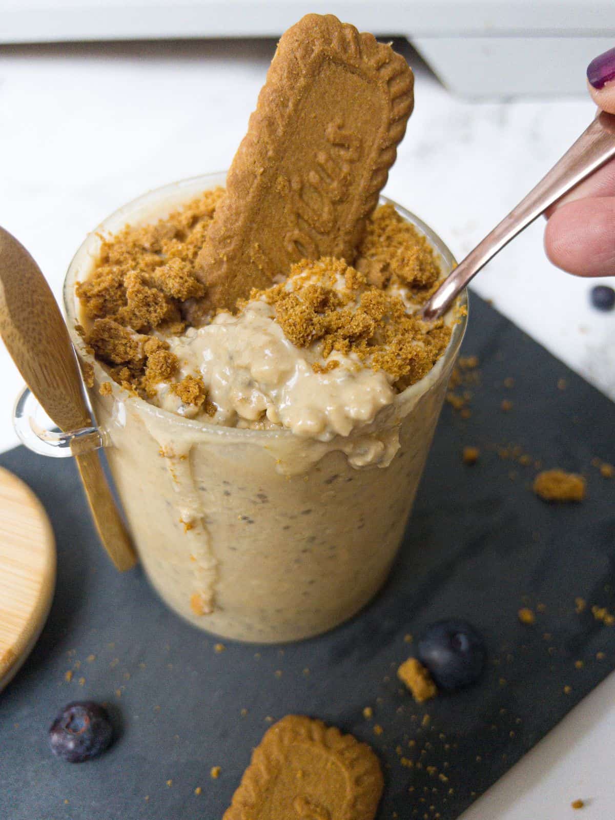 Overnight oats in a glass jar, with a spoon being dipped into it,topped with crumbled Biscoff biscuits and a whole biscoff biscuit. Jar sitting on a gre board with crumbs and a biscuit scattered around.