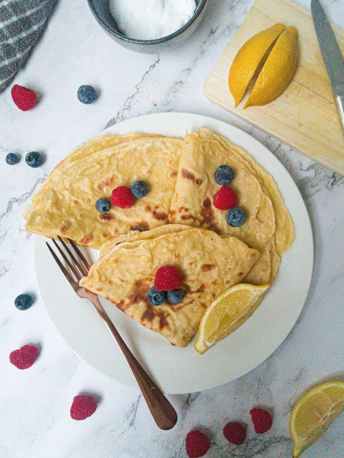 Three crepes on a white plate garnished with raspberries & blueberries. there is a fork on the plate and sliced lemon. In background there is a lemon on a chopping board and a bowl of sugar.
