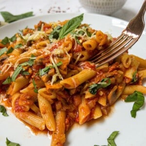 White plate filled with penne pasta in a tomato sauce, garnished with basil and cheese. A fork is picking up a piece of pasta.