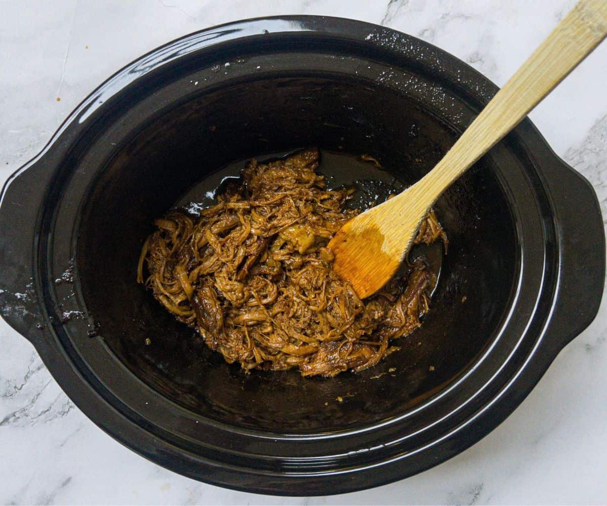 Shredded cooked beef brisket in a slow cooker with a wooden spoon in it.
