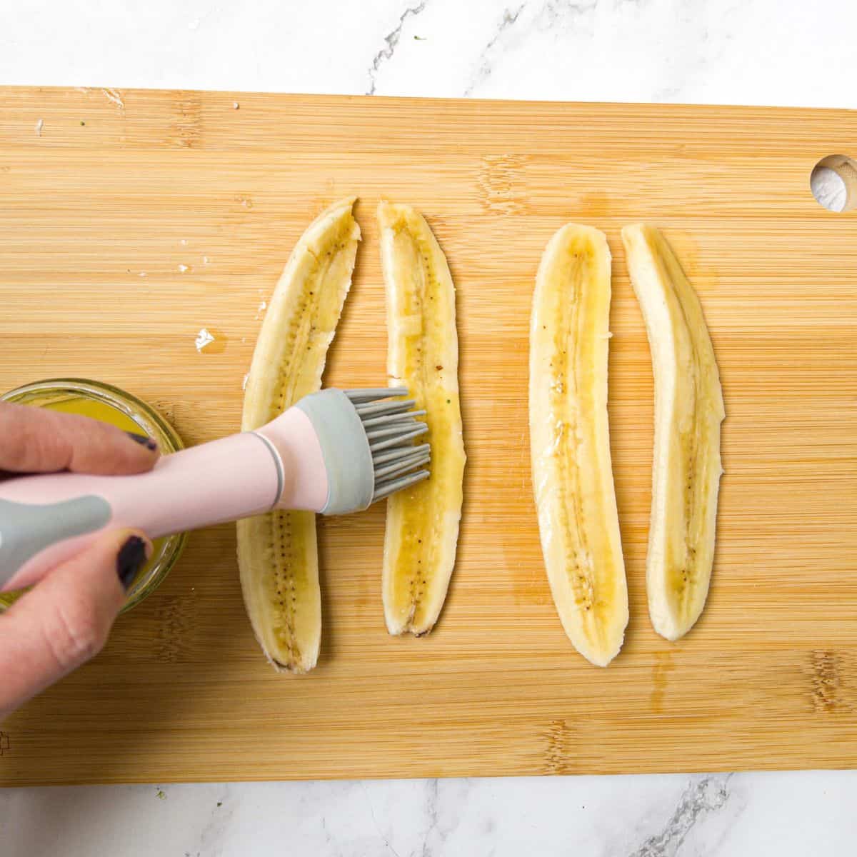 Two bananas sliced lengthways being basted with butter using a pastry brush.