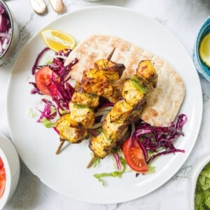 Two chicken shish kebabs on sticks laying on top of salad and pitta bread on a white plate.