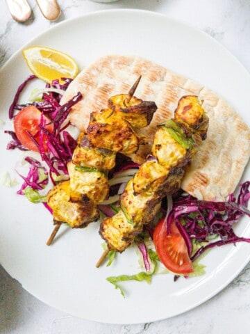 Two chicken shish kebabs on sticks laying on top of salad and pitta bread on a white plate.