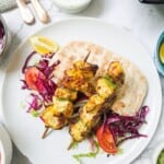Two chicken shish kebabs on sticks laying on top of salad and pitta bread on a white plate. With a bowl of red cabbage and a pot of garlic sauce in the background.
