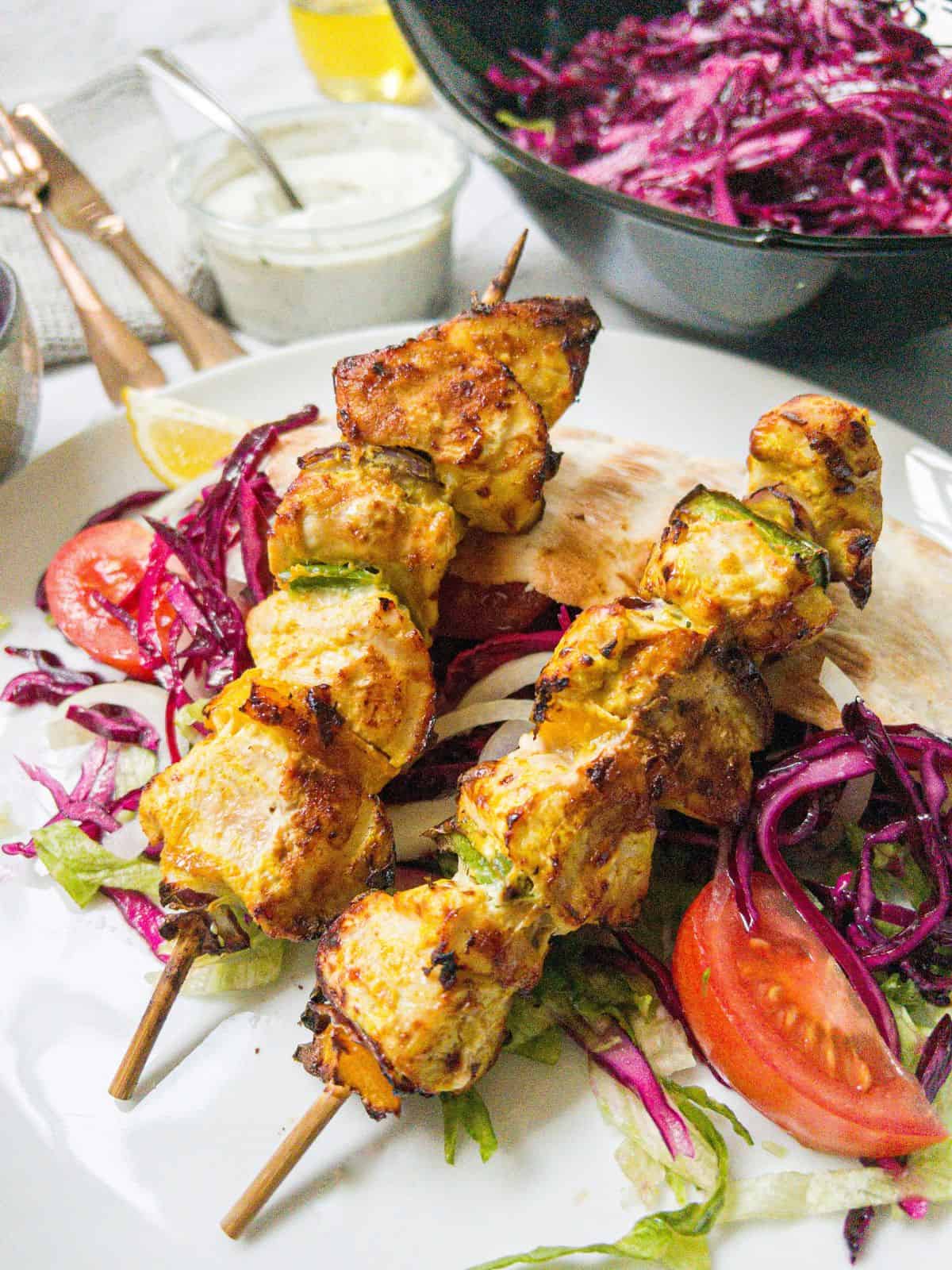 Two chicken shish kebabs on sticks laying on top of salad and pitta bread on a white plate. With a bowl of red cabbage & pot of garlic sauce in the background.