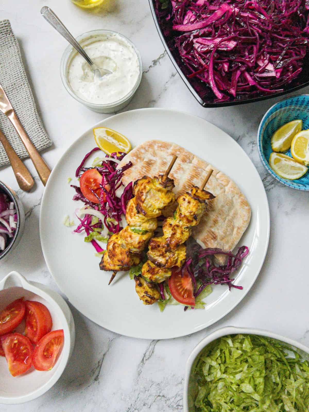 Two chicken shish kebabs on sticks laying on top of salad and pitta bread on a white plate. With a bowl of red cabbage, a pot of garlic sauce, some lemon wedges, tomatoes and lettuce in the background.