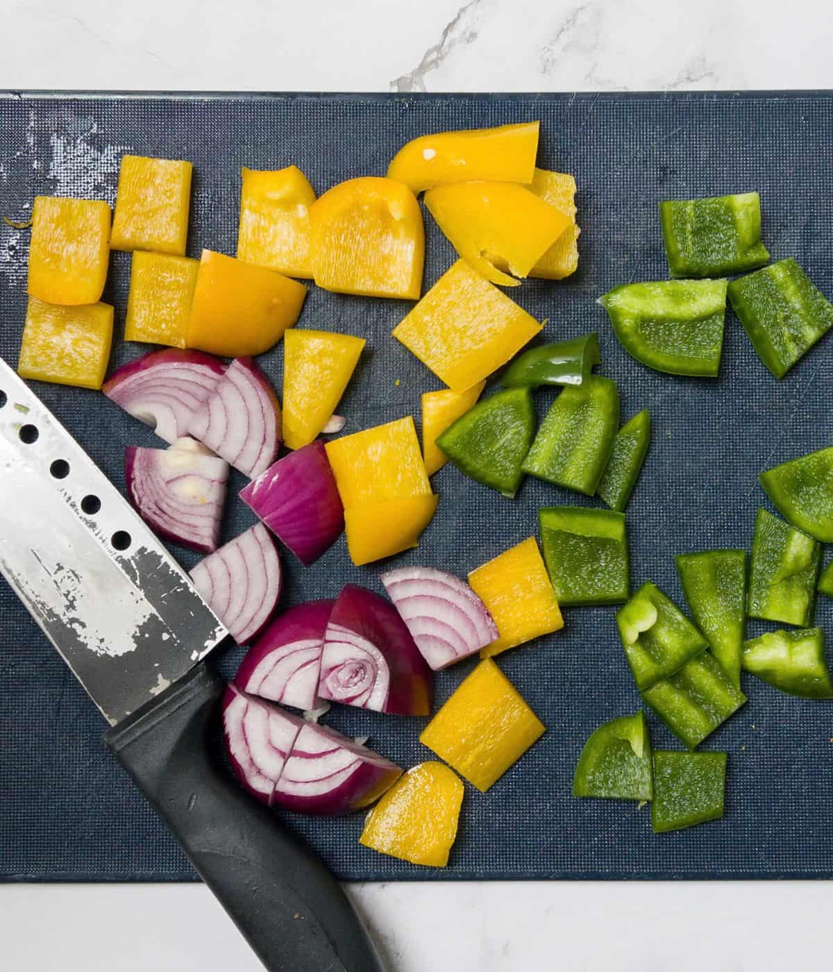 Chopped green and yellow peppers and red onion on a blue chopping board with a knife.