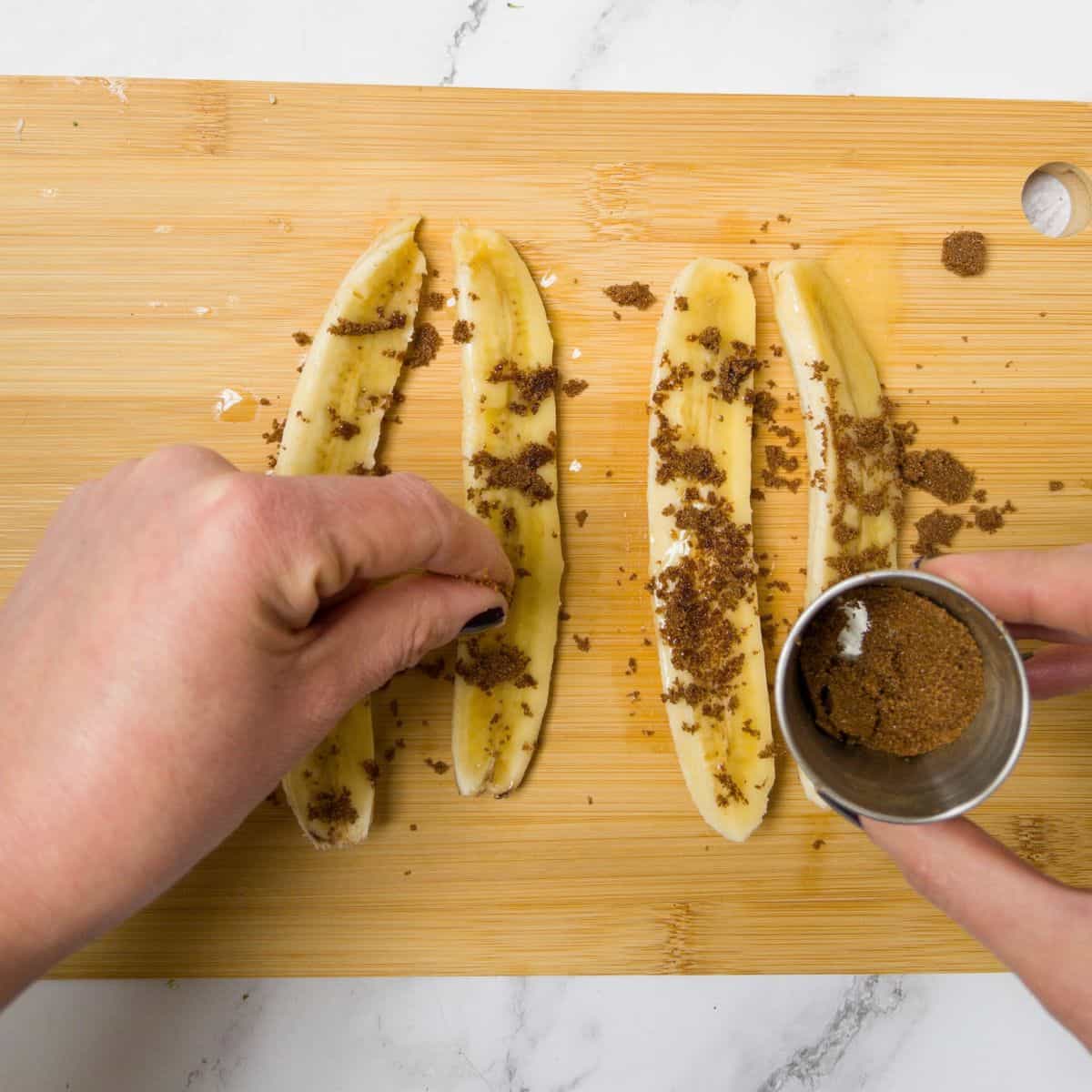 Four sliced bananas on a chopping board with someone sprinkling brown sugar on them.